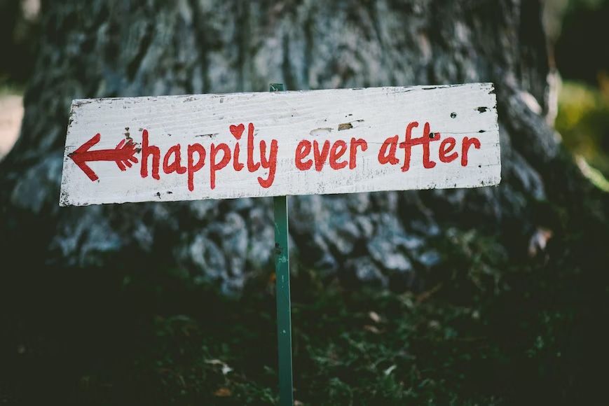 A short pole with a sign posted in front of a tree that says "happily ever after." with an arrow pointing to the left.