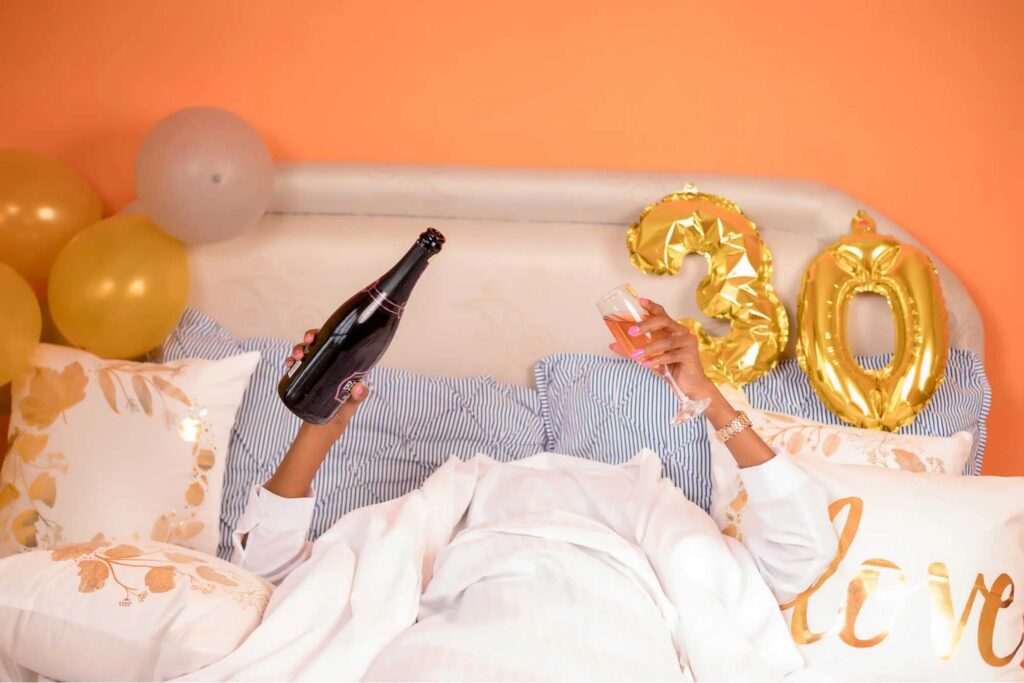 A man hiding in his comforter while holding a wine and wine glass during his early morning 30th birthday.