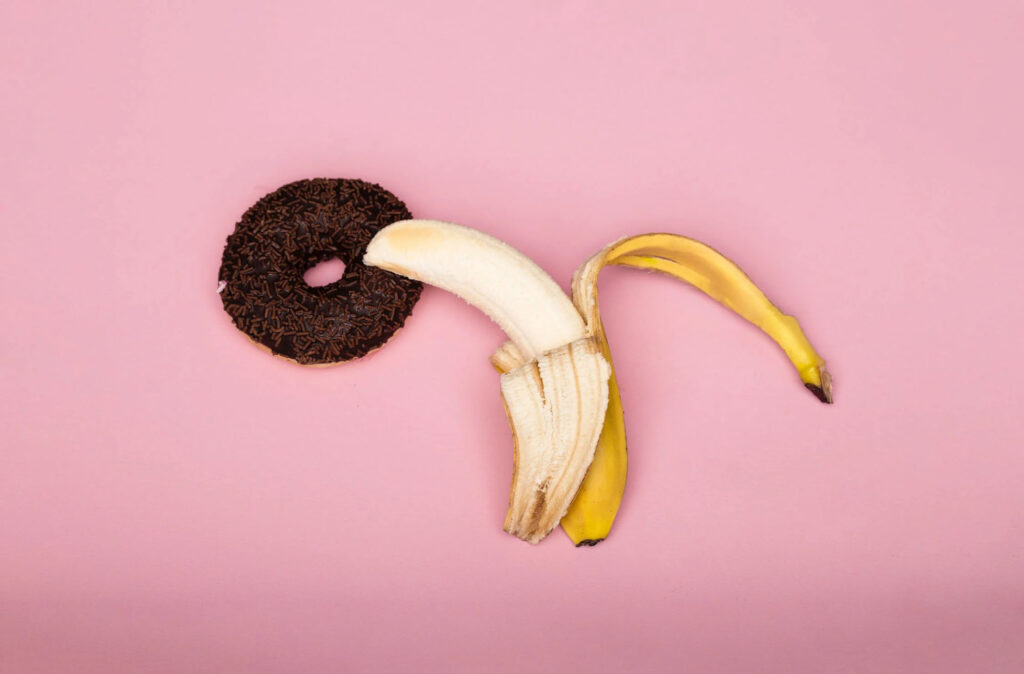 A banana beside a chocolate flavored donut on a pink background.