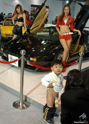 Sexy sales ladies posing near a black supercar while a kid in front of them pees in a bottle.