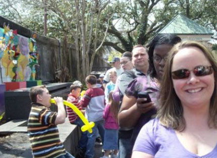 A woman taking a selfie while falling in line when a kid behind her puts a balloon animal in his mouth.