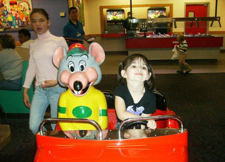 A kid rides on a cart with a Chuck E Cheese mouse while another kid from behind walks by with pants down.