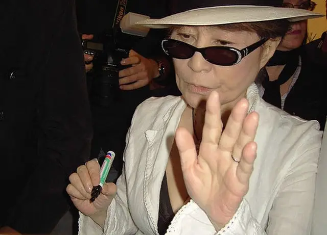 Yoko Ono in white beach hat, dress, and dark shades holding a pen for autograph signing.