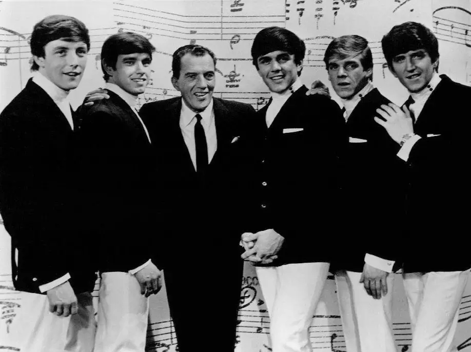 1964 back and white photo of The Dave Clark Five (five young men) with Ed Sullivan at the center from an appearance on Sullivan's show.