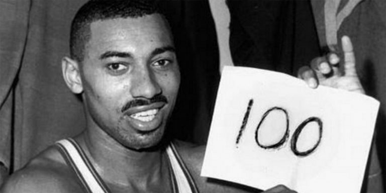 A black and white photo of Wilt Chamberlain holding a paper with a number 100 written on it.