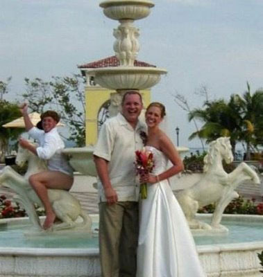 Newly-wed couples posing in front of a fountain when a half-naked guy on the background rides on a horse statue.