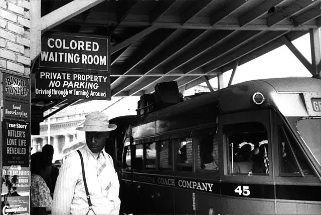 A black and white photo of a colored man waiting at the train station under the sign, "Colored Waiting Room".