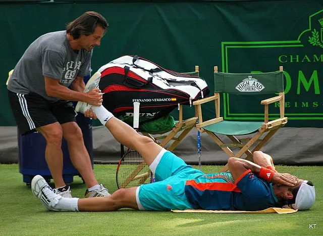 A coach pulling the leg of a tennis player while he lies on the ground due to a sprain.