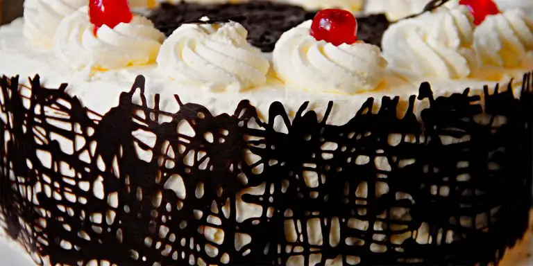 A cake with chocolate drizzle on the side, white cream on top with icing and a cherry over it.