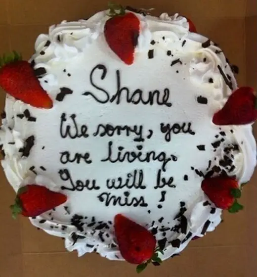 An apology cake with white cream, strawberries on top, and a message that says, "Shane. We sorry, you are living. You will be miss!"